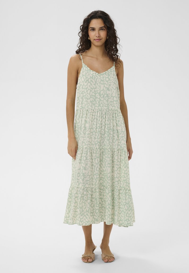 Femme portant une robe midi sans manches, verte clair à volants avec un motif abstrait, et des sandales beiges à bouts ouverts, debout devant un fond uni.