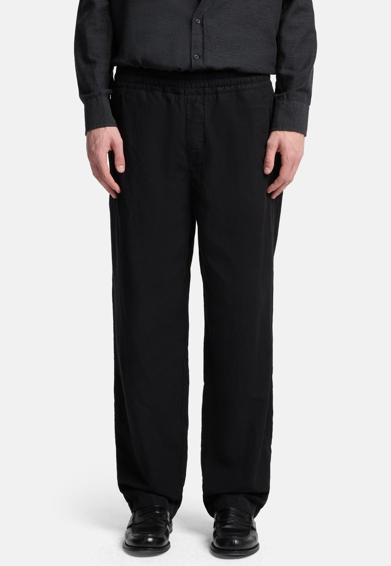 Schwarze Hose mit entspannter Passform, elastischem Bund und glatter Textur, kombiniert mit schwarzen Lederschuhen.