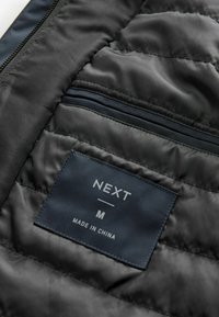 Jachetă puf neagră cu căptușeală cu textură matlasată, buzunar cu fermoar și etichetă din material cu "NEXT," mărimea "M," și "FABRICAT ÎN CHINA" imprimat.