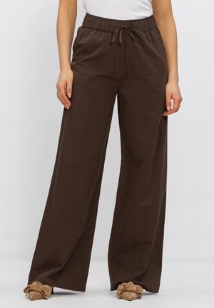 Bruine wijde broek met elastische tailleband en trekkoord, gecombineerd met beige platte schoenen met een strikaccent en een wit topje.