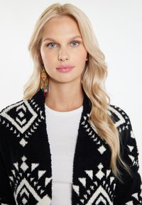 Svart och vit mönstrad cardigan i mjukt material med geometriska mönster, bärs över en vit skjorta, med långt, vågigt blont hår.