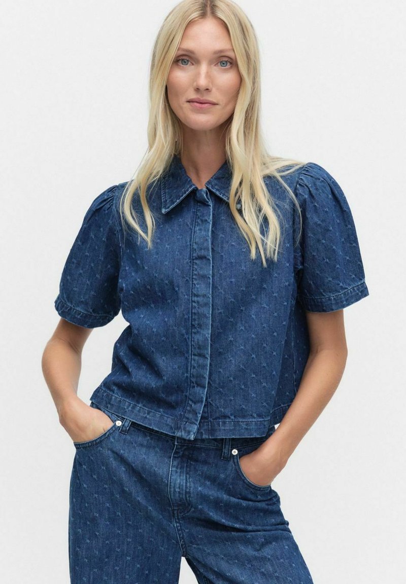 Blonde vrouw met blauwe ogen, gekleed in een bijpassende donkerblauwe gestructureerde spijkerblouse met pofmouwen en hoge taille jeans, met haar handen in haar zakken.