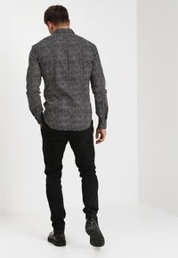 Camisa negra de manga larga con un patrón de puntos blancos, ajuste slim, combinada con pantalones negros y botas negras, vista desde la parte de atrás.