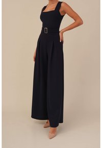 Mujer con un mono negro de pierna ancha y escote cuadrado, cinturón y tacones de punta desnuda frente a un fondo beige.