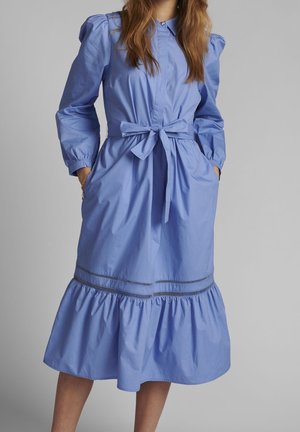 Femme portant une robe bleue mi-longue avec des manches longues bouffantes, une ceinture nouée à la taille et un ourlet à volants, debout les mains dans les poches.