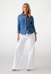 Kobieta stoi w dopasowanej niebieskiej koszuli dżinsowej, szerokich białych spodniach i srebrnych sandałach na obcasie, na tle jednolitego jasnego tła.