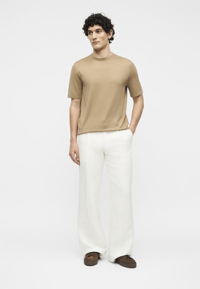 Άνδρας με σγουρά μαλλιά που φοράει μια μπεζ κοντομάνικη πουκαμίσα, λευκό παντελόνι με φαρδιά μπατζάκια και καφέ παπούτσια, στέκεται με το ένα χέρι στην τσέπη.