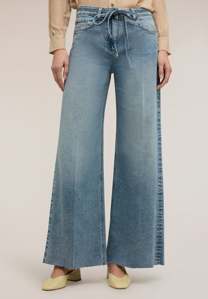 Breitbeinige hellblaue Jeans aus Denim mit ausgefransten Säumen und einem gebundenen Gürtel, kombiniert mit einer beigen Bluse und cremefarbenen Schuhen mit Blockabsatz.