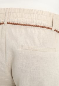 Béžové lněné kalhoty s elastickým pasem a pleteným hnědým detailem, mají jedno zadní kapsu. Texturovaná tkanina s jemným leskem.