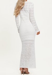 Longue robe blanche en dentelle avec manches longues, silhouette ajustée, motifs complexes et un ourlet subtilement festonné, associée à des escarpins beige. 