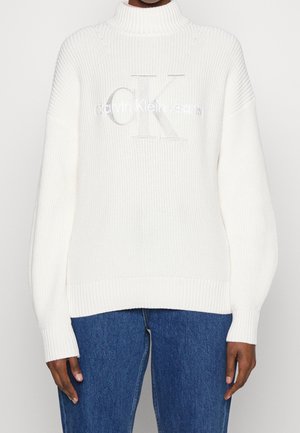 Witte geribbelde gebreide trui met hoge hals, voorzien van het 'Calvin Klein Jeans' logo en lange mouwen. Gepaard met blauwe denim jeans.