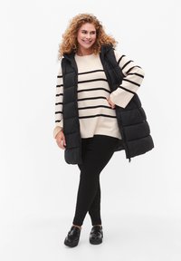 Svart pufferväst över en krämfärgad topp med svarta ränder, ihop med svarta leggings och glänsande loafers. Lockigt hår, står framför en neutral bakgrund.