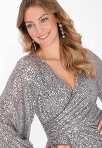 Hopeinen paljettinen kietaisumekko, jossa pitkät, laskeutuvat hihat. Teksturoidusta kankaasta, v-pääntiellä ja kerätyllä vyötäröllä, korostaen laskeutuvaa siluettia.