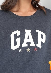 Harmaa huppari, jossa on näyttävästi valkoisilla kirjaimilla "GAP" ja alla kolme tähteä punaisena, kullanvärisenä ja sinisenä.