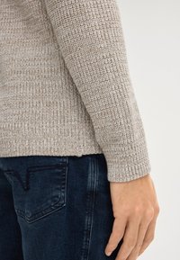 Beżowy sweter wykonany na drutach z teksturowanym wzorem, z długimi rękawami i lekko prążkowanym dołem. Połączony z ciemnoniebieskimi dżinsami.