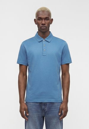 Ung mand med kort blondt hår iført en blå polo med korte ærmer og blå jeans, står mod en ensfarvet, lys baggrund.