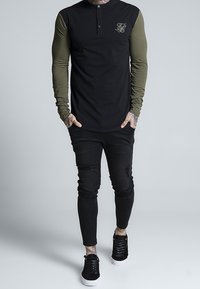 Black long-sleeve shirt with olive green sleeves, featuring a buttoned neckline and logo. Paired with black ripped skinny jeans and black sneakers.