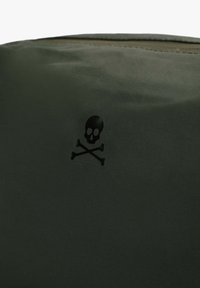 Bolsa de tela verde con un emblema negro de calavera y huesos cruzados. La superficie tiene una textura suave con costuras a lo largo de los bordes.