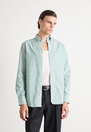 Camisa de botonadura en azul verde claro con un bolsillo, hecha de un tejido texturizado, combinada con una camiseta blanca de tirantes y pantalones negros.