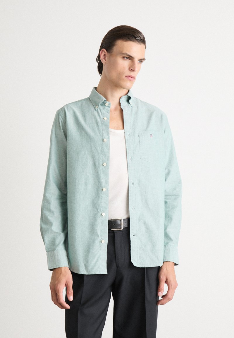 Camicia azzurro-verde chiaro con bottoni e taschino, realizzata in un tessuto texturizzato, abbinata a una canottiera bianca e pantaloni neri.