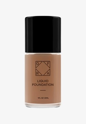 Fond de teint liquide dans une bouteille en verre transparent, avec un capuchon cylindrique noir. L'étiquette comprend le logo de la marque et le nom du produit en texte noir.