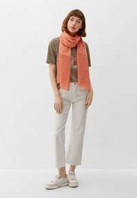 Koraal kleurige sjaal met oogjespatroon om de hals, gecombineerd met een grijs T-shirt met korte mouwen, lichtbeige cropped broeken en witte sneakers.