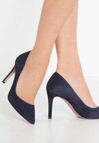 Mörkblå mocka högklackade skor med spetsig tå och en 10 cm hög stilettklack. Slät textur och minimalistisk design utan dekorationer.