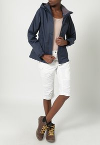 Granatowa wodoodporna kurtka z kapturem, beżowa bluzka, białe krótkie spodnie bojówki i brązowe buty turystyczne. Teksturowany design z widocznymi szwami i zamkami.