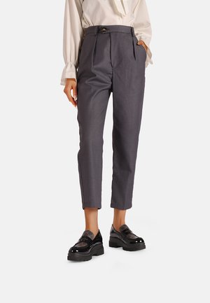 Pantaloni grigi a vita alta con vestibilità sartoriale, dotati di passanti per la cintura, tasche frontali e una piega lungo la gamba. Mocassini neri con suola spessa.