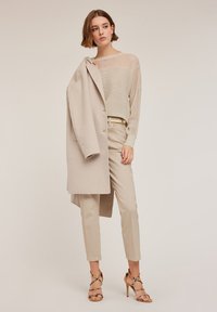 Beige ullkappa över en ljus randig stickad topp, med ljusa byxor och beiga högklackade sandaler med remmar, vilket skapar en neutral palett.