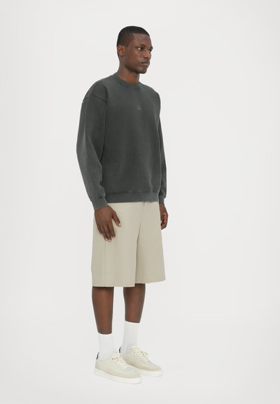 Mørkegrå sweatshirt med ribbestrik ved håndleddene, parret med lysebeige shorts. Modellen har cremefarvede sportssko og hvide ankelsokker på. Klar baggrund.