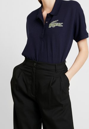 Vrouw draagt een marineblauw poloshirt met een groen geborduurd logo en zwarte plissébroek met hoge taille, hand in de zak.