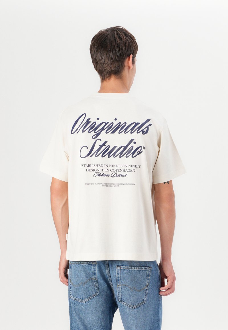 T-shirt en coton couleur crème avec un grand texte bleu marine au dos. Le design comprend des détails sur son origine et son but.