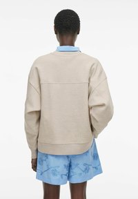 Beżowy sweter z luźnym krojem i opadającymi ramionami, z prostym dołem. W połączeniu z niebieskimi szortami z haftem.