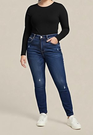 Personne portant un haut noir à manches longues, un jean bleu foncé déchiré et des baskets blanches, une main dans la poche sur un fond uni.