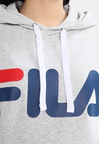 Sudadera gris con bolsillo frontal, cordones blancos y un gran logotipo de FILA en rojo y azul marino. La tela parece suave y con textura.