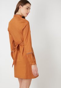 Vestido color óxido con cintura ajustada, mangas plisadas y detalle de lazo en la espalda. La tela suave y el diseño del cuello completan el look.
