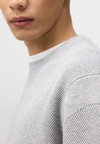 Lichtgrijze gebreide trui met een gestructureerd patroon, ronde halslijn en een gelaagde witte shirt zichtbaar eronder. Close-up van de stof en het halsgebied.