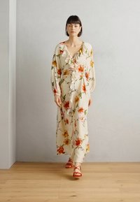 Abito maxi floreale in crema, con scollo arricciato e maniche lunghe. Fiori rossi e gialli vivaci con foglie verdi. Indossato con sandali rossi con cinturini.