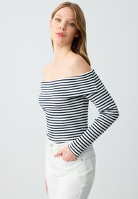 Schulterfreies, geripptes Oberteil in Weiß mit navyblauen Streifen. Lange Ärmel, taillierte Passform mit strukturierter Optik. Kombiniert mit weißen Hosen.