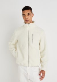 Casaco de fleece em off-white com gola alta, fecho éclair frontal e bolso no peito com fecho éclair; apresenta um tecido suave e texturizado.