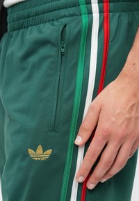 Πράσινα αθλητικά παντελόνια jogging με υφή, που διαθέτουν τσέπη με φ ερμουάρ, λευκές, πράσινες και κόκκινες ρίγες στο πλάι και χρυσό λογότυπο.