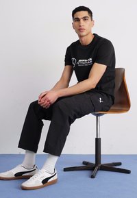 Zwart T-shirt met wit logo, zwarte cargobroek, witte sokken en witte sneakers met zwarte accenten. Model zittend op een houten stoel.