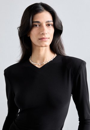 Haut noir à manches longues avec un décolleté en V. Texture lisse, coupe ajustée. Un collier de perles notable avec des perles de tailles variées rehausse le look.