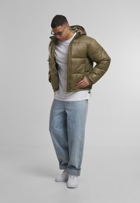 Olíva zöld puffdzseki kapucnival, steppelt részekkel és cipzáros előlappal. Világoskék farmernadrággal és fehér sneaker-zel párosítva.