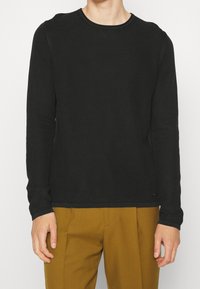 Homme portant un pull noir texturé à manches longues et col rond, ainsi qu'un pantalon plissé marron moutarde, debout devant un fond uni.