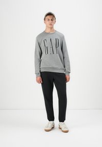 Sudadera gris con el logo "GAP", cuello redondo y puños acanalados, combinada con pantalones negros ajustados y zapatillas crema con suelas marrones.