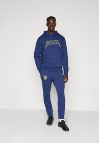 Mand stående iført en blå hættetrøje og matchende sweatpants med gule Atletico Madrid-logoer og sorte sneakers foran en ensfarvet baggrund.