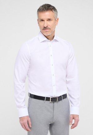 Weißes Langarmhemd mit Button-Down-Kragen und Frontblende, kombiniert mit grauen Hosen und einem schwarzen Ledergürtel. Glatte Stofftextur.