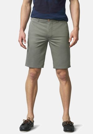 Man staat en draagt olijfgroene knielange korte broek, een donkerblauw overhemd en zwarte leren bootschoenen met bruine veters.